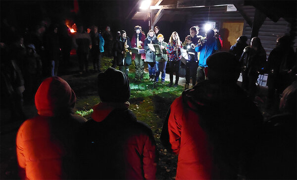 Studierende musizieren vor einer Waldhütte bei Nacht. Zuschauer stehen drumherum.