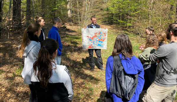 Prof. Bertil Burian steht mit Studierenden im Wald und zeigt eine Karte vom Rammert. Neben grundlegenden Infos zur Geographie sowie den verschiedenen Baumarten, teilt Herr Burian auch vertiefendes forstwirtschaftliches Wissen mit. 