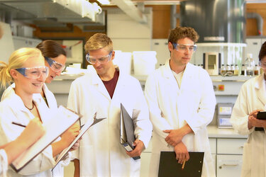 Eine Gruppe Studierende steht mit weißen Laborkitteln und Schutzbrillen im Labor der Hochschule Rottenburg und machen sich Notizen