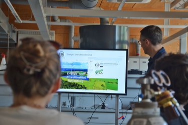 Studierende sitzen vor einem Monitor und hören dem Dozenten zu