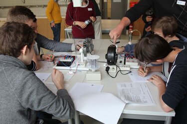 5 Schüler sitzen gemeinsam an einem Tisch im Labor und machen sich Notizen zu einem Versuch.