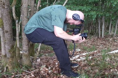 Ein Kameramann lehnt in der Hocke an einem Baum und schaut durch seine Kamera und filmt