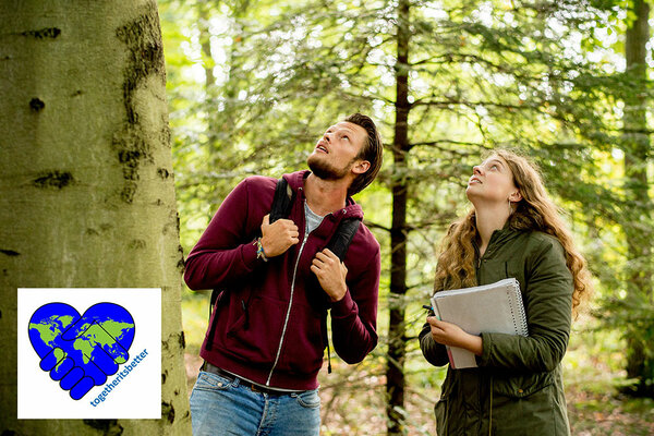 Eine Studentin und ein Student stehen im Wald und blicken in das Kronendach. Die Studentin hat einen Schreibblock in der Hand und macht Notizen. 