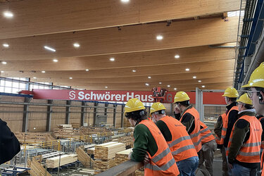 Studierende mit orangenen Warnwesten und gelben Helmen schauen von oben in die Produktionshalle