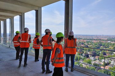 Studiernede stehen mit orangenen Warnwesten und Helmen auf der Baustelle