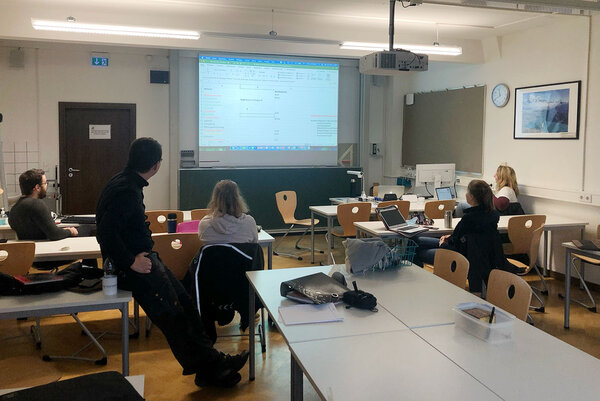 Studierende besprechen sich gemeinsam in einem Seminarraum und schauen auf eine Präsentation auf der Leinwand