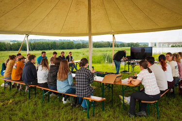 Studierende sitzen bei einem Vortrag auf Bierbänken unter einem großen Sonnenschutz im Freien und schauen auf einen aufgebauten Monitor