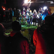 Studierende musizieren vor einer Waldhütte bei Nacht. Zuschauer stehen drumherum.