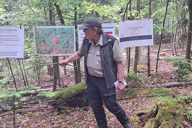 Im Wald sind Plakate und Karten mit Wäscheklammern an einer Schnur befestigt. Ein Referent steht davor und erklärt sie mithilfe eines Zeigestocks.