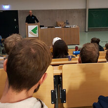 Studierende sitzen in der Aula. Blick nach vorne zum Referenten.