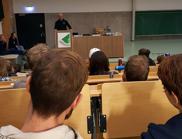 Studierende sitzen in der Aula. Blick nach vorne zum Referenten.