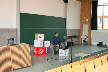 Prof. Abele sitzt auf der Bühne der Aula und referiert.