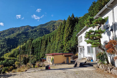 Blick auf ein weißes Haus. Im Hintergrund bewaldete Berge.