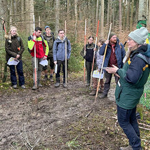 Studierende stehen mit Fluchtstäben in der Hand auf einem Waldweg und höhren der Referentin zu