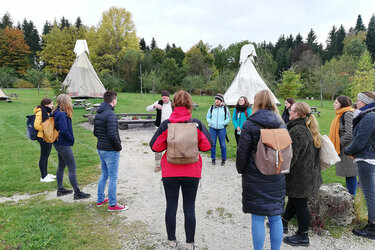 Studierende stehen vor Tipis auf dem Hofgut Hopfenburg und hören dem Referenten zu