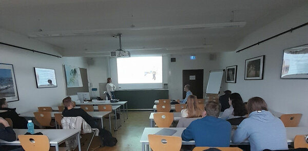 Blick von hinten in einem Seminarraum nach vorne. Vorne steht die Referentin und Studierende sitzen an Tischen. 