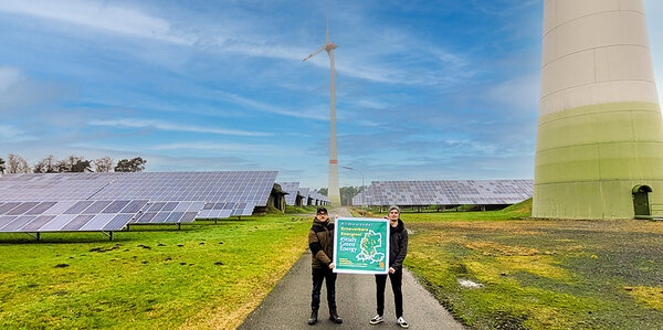 Studierende halten das Plakat "Klimawandel - Erneuerbare Energien - 'Study green energy" in die Kamera. Im Hintergrund sin Windräder und Photovoltaikanlagen zu sehen