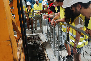 Auf einer Baustelle stehen Lernende in gelben Warnwesten und weißen Helmen und lauschen dem Vortragenden