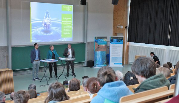 Die Referenten stehen auf der Bühne der Aula.