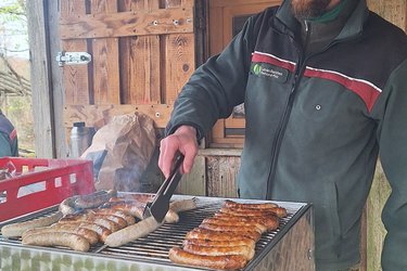 Ein Förster steht hinter einem Grill und grillt Würstchen für die Gäste