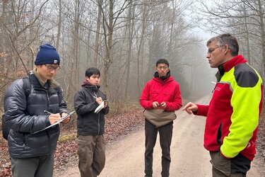 Prof. Beimgraben steht mit einer Gruppe von Gästen auf einem Waldweg und gibt Erklärungen.