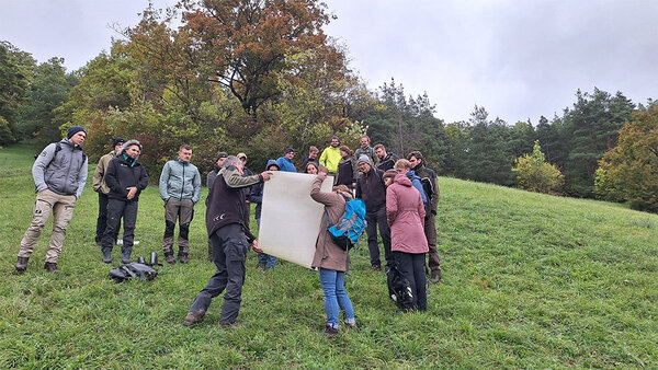 Studierende stehen auf einer Wiese und betrachten eine große Karte, die der Referent mit einer Hilfe in die Luft hält.
