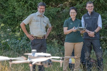 Drohenpilotin läösst eine Drohne über dem Boden schweben. Zwei Personen stehen neben der Pilotin und schauen zu