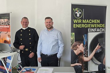 Zwei Schornsteinfeger stehen an einem Infostand und lächeln in die Kamera