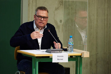 Steffen Rathke sitzt an einem Tisch mit Tischmikrofon und Plexiglaswand zwischen dem Nachbartisch