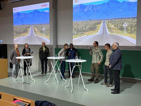 Die Referenten stehen auf der Bühne der Aula an Stehtischen