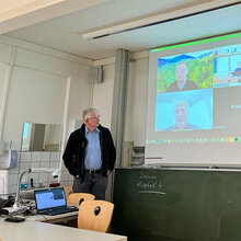 Prof. Schurr steht vor der an die Wand projizierte Zoom-Sitzung