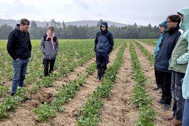 Studierende stehen auf einer landwirtschaftlichen Fläche und hören dem Dozenten zu