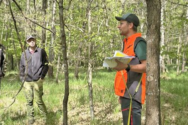 Referent mit Unterlagen in der Hand steht im Wald und referiert vor den Studierenden. Der Referent hat eine orangene Weste an