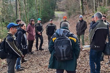 Studierende stehen auf einem Waldweg und hören dem Referenten zu.