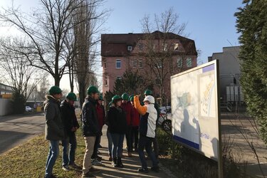 Die Exkursionsteilnehmer stehen in einer Gruppe zusammen und tragen grüne Helme. Die Exkursionsleiterin erläutert das Müllheizkraftwerk an einer Infotafel.