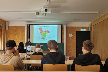 Blick von hinten in einen Hörsaal. Vorne steht Prof. Poetsch vor den Schülern und referiert. An die Leinwand ist eine Präsentation projiziert.