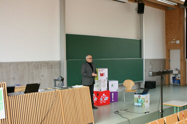 Prof. Kaiser steht auf der Bühne der Aula und referiert. 