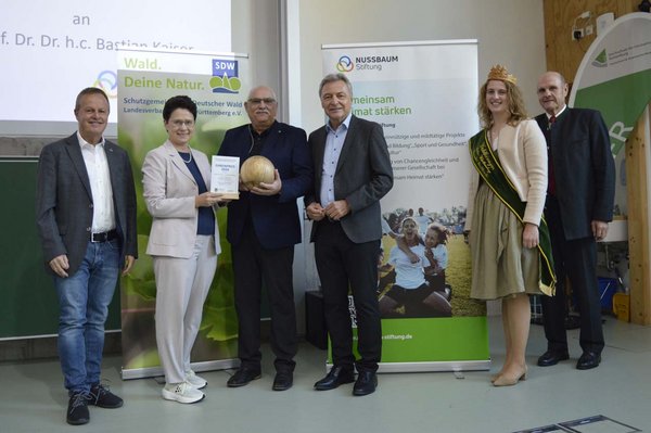 Prof. Kaiser bekommt den Preis auf der Bühne überreicht. v.l.n.r.: Ulrich Kienzler (SDW BW Karlsruhe), Marion Gentges MdL (Ministerin für Ländlichen Raum, Landwirtschaft und Heimat), Preisträger Prof. Dr. Bastian Kaiser, Peter Jahn (NUSSBAUM Stiftung), Franziska Waselikowski (Waldkönigin Baden-Wüttemberg) und Karl-Wilhelm Röhm (Landesehrenvorsitzender SDW BW) bei der Verleihung des SDW Ehrenpreises 2026. (Foto: SDW BW)