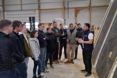 Studierende stehen in der Werkshalle und hören dem Referenten zu