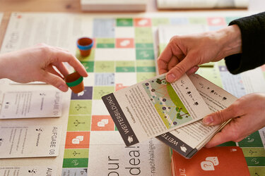 Blick auf das Brettspiel Pitch Your Green Idea. Über dem Brettspiel sind Hände von zwei Spielenden zu sehen. Eine Person teilt Karten aus und die andere zieht eine Spielfigur