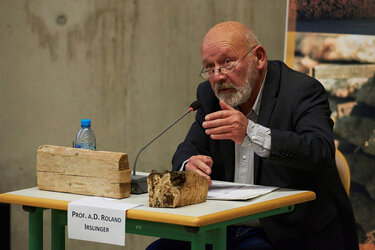 Prof. a.D. Roland Irslinger sitzt an einem Tisch mit Tischmikrofon und Plexiglaswand zwischen dem Nachbartisch