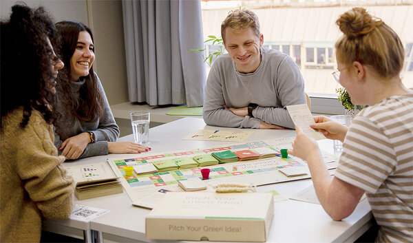 Drei Studentinnen und ein Student sitzen zusammen um einen Tisch und spielen zusammen das Brettspiel Pitch Your Green Idea