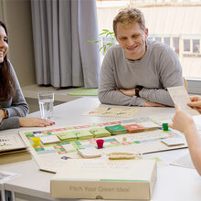Drei Studentinnen und ein Student sitzen zusammen um einen Tisch und spielen zusammen das Brettspiel Pitch Your Green Idea