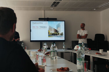 Studierende in einem Konferenzraum. Der Blick ist auf einen Bildschirm mit einer Präsentation und einen Referenten gerichtet.