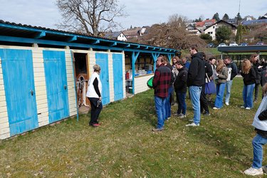 Studierende stehen in dem Naturbad im Kreis vor den Umkleidekabinen und hören dem Referenten zu