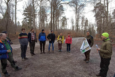 Die Teilnehmer stehen im Wald um die Referenten. Die Referenten halten eine Karte in der Hand.