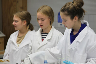 3 Schülerinnen stehen gemeinsam an einem Tisch im Labor und experimentieren mit Biomasse