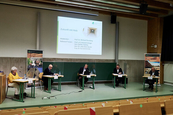 Blick auf das Podium der Aula mit den Referenten. Die Referenten sitzen an einzelnen Tischen mit Plexiglaswänden dazwischen