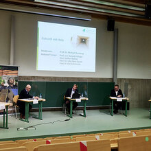 Blick auf das Podium der Aula mit den Referenten. Die Referenten sitzen an einzelnen Tischen mit Plexiglaswänden dazwischen