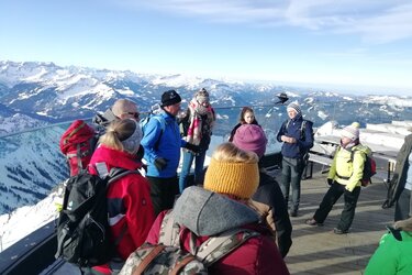 Herr Joachim unterhält sich mit der Teilnehmergruppe. Im Hintergrund sieht man die verschneiten Berggipfel